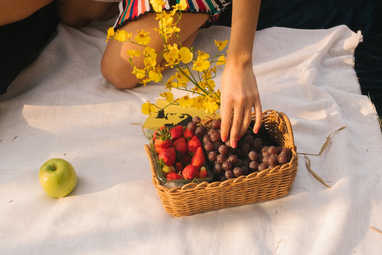 Home A vibrant picnic setup with strawberries, grapes, an apple, and yellow flowers in a wicker basket.